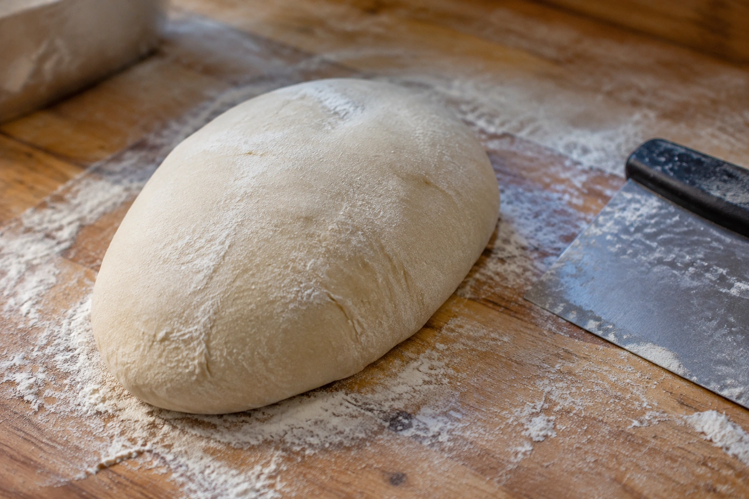 Oval-shaped bread dough on a floured bench beside a dough scraper
